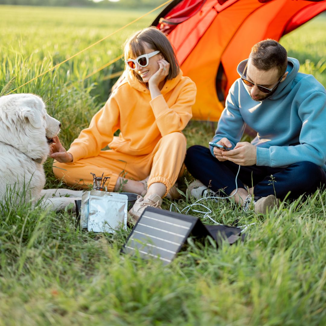 Two people and a dog sitting in a grassy field with a solar panel and orange tent in the background.