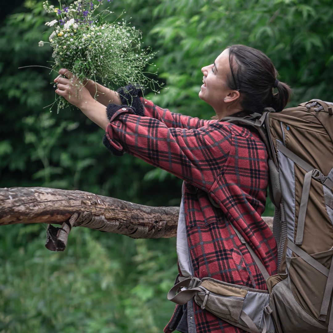Person in a red plaid shirt with a backpack holding flowers in a forest setting