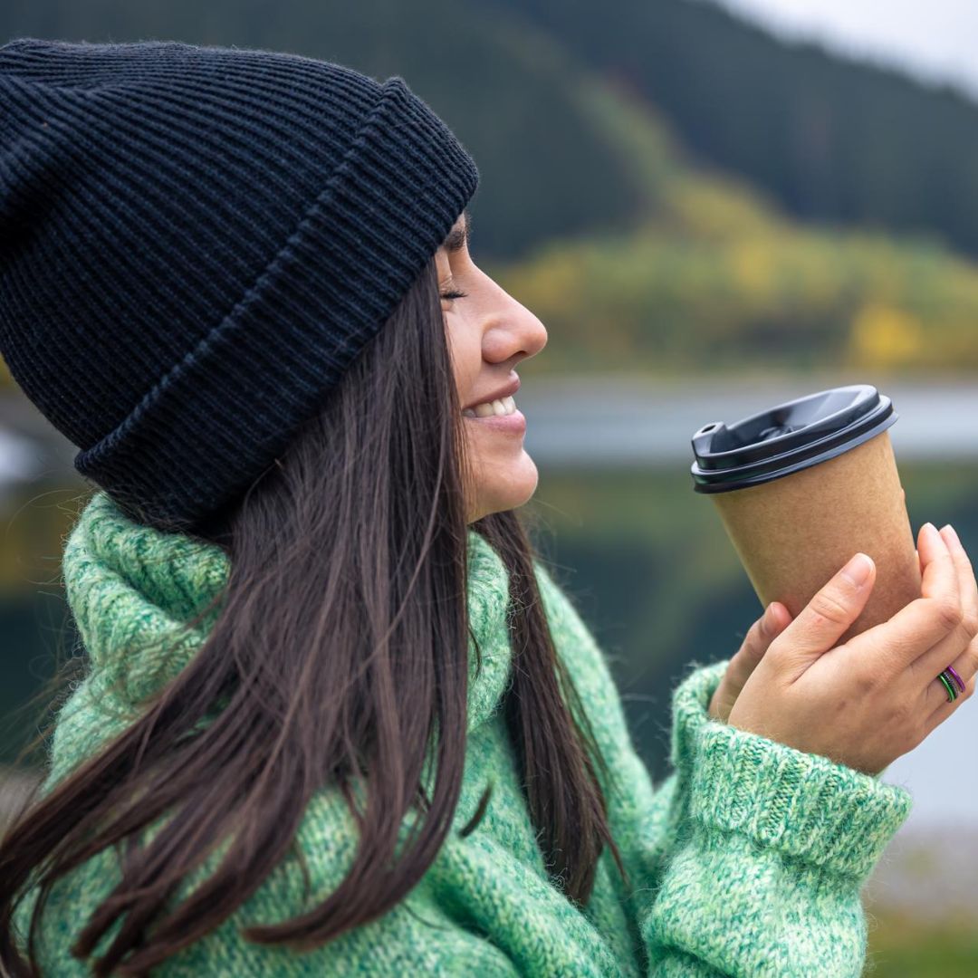 Woman in green sweater and black beanie holding a coffee cup outdoors with a scenic background